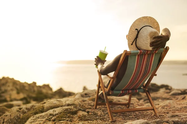 非洲女孩的帽子和饮料在她的手上面对大海的日光浴 — 图库照片