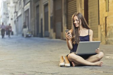 Şehir merkezinde sokak üzerinde bir bilgisayar ve bir akıllı telefon kullanan kız