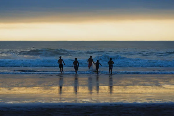 Siluetas de cuatro chicos (jóvenes) en la playa corriendo hacia el oleaje al atardecer . — Foto de Stock