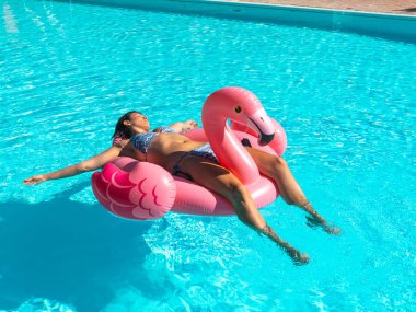 Genç bir kadın şişme pembe flamingo oyuncağında havuzda dinleniyor.