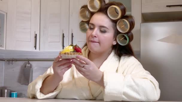 Plus-size girl, dressed in a bathrobe, curlers on her head, looks at the cake with desire, not daring to start eating it — Stock video