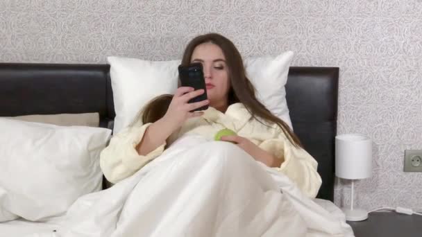 Tamaño más joven mujer acostada en la cama, mensajería en el teléfono inteligente, cambiar los canales de televisión — Vídeo de stock