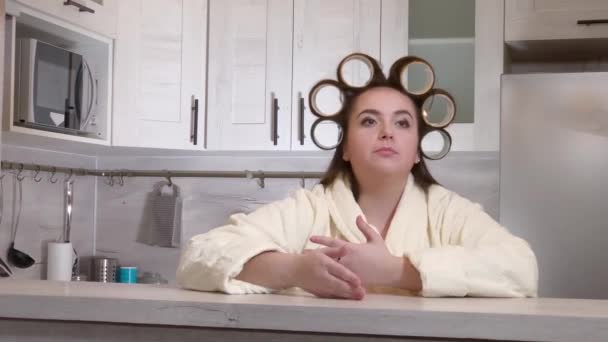 Plus-size girl dressed in a bathrobe, curlers on her head, emotionally speaks to camera — Stock Video