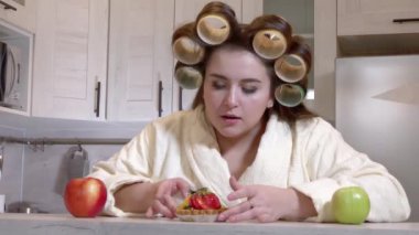 Plus-size girl, dressed in a bathrobe, curlers on her head, looks at the cake with desire, not daring to start eating it