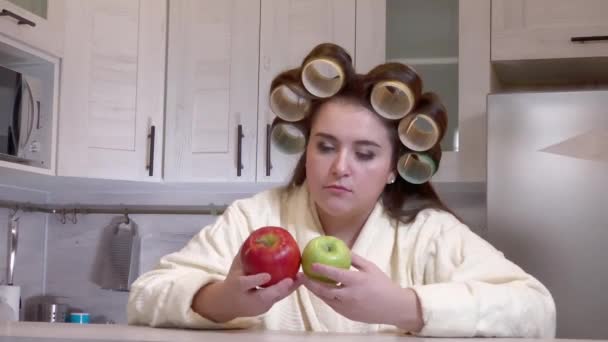 Plus-size girl, dressed in a bathrobe, curlers on her head, looks at apples, she does not want to eat them — Stock video