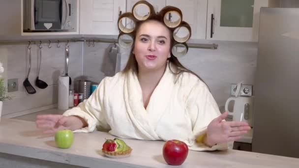 Plus-size meisje met krulspelden op haar hoofd gekleed in een badjas is het opnemen van een video blog — Stockvideo