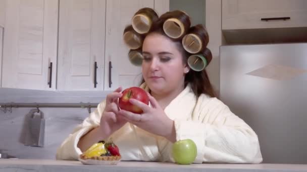 Плюс-размер девушка, одетая в халат, бигуди на голове, смотрит на яблоки, она не хочет есть их — стоковое видео