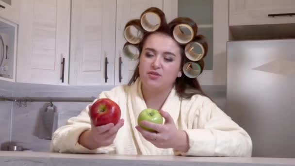 Plus-size girl, dressed in a bathrobe, curlers on her head, looks at apples, she does not want to eat them — Stock video