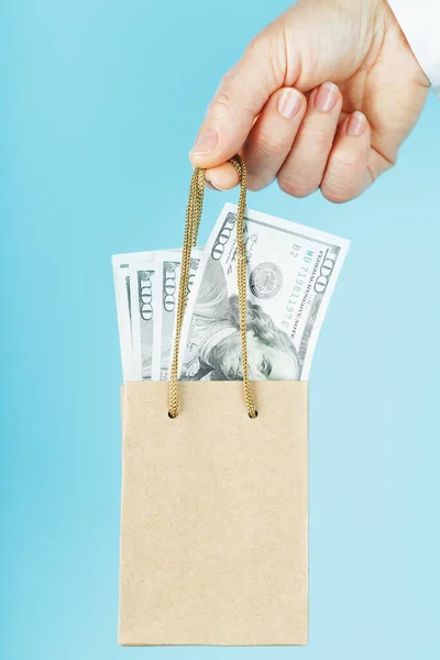 A small paper bag for financial aid and support made of paper at arm\'s length with US dollars on a blue background. The concept of negligible financial support in business, the population and entrepreneurs. The concept of financial assistance.Dollar