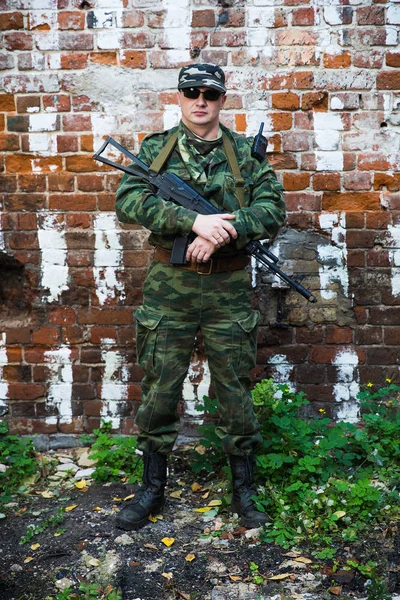 Soldado o miliciano en camuflaje con rifle de asalto AK-74 permanecer cerca de la pared de ladrillo — Foto de Stock