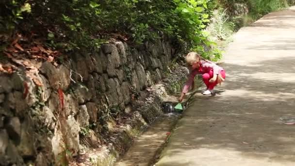 Chica corriendo por el camino con un barco de papel en la mano. Se detiene cerca del arroyo y lo corre en la natación — Vídeo de stock