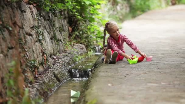 Девушка сидит у ручья и запускает много бумажных лодок на воду и машет рукой — стоковое видео