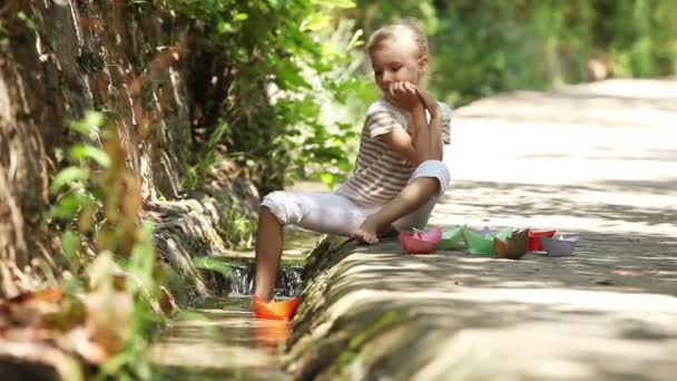 Niño sentado cerca del arroyo. Chica está lanzando un barco de juguete flotando a lo largo del arroyo — Vídeos de Stock