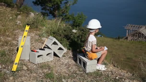 A criança pequena é a construtora. Menina no capacete de construção e com óculos de sol. Ela está escrevendo no caderno — Vídeo de Stock