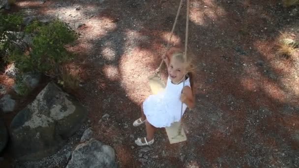 Menina feliz em vestido branco com cabelo loiro em um balanço. Ela está feliz e gosta. Criança sorrindo para a câmera e girando — Vídeo de Stock