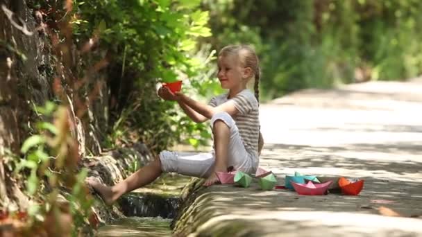 Ragazza seduta accanto a un ruscello e che lancia un sacco di barche di carta sull'acqua e mano ondulata — Video Stock