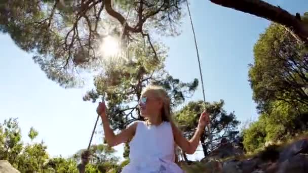 Menina bonito em vestido branco com óculos de sol em um balanço. Ela está feliz e rindo da câmera — Vídeo de Stock