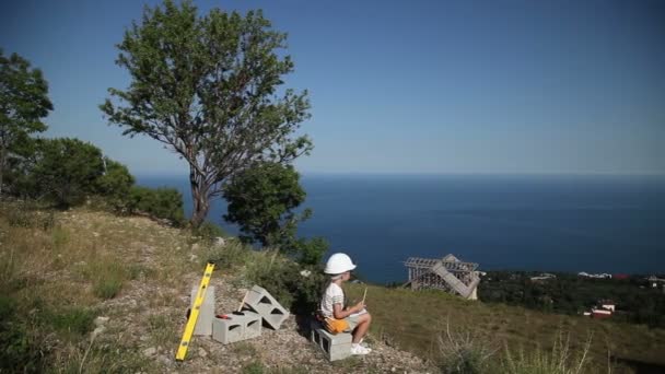 A criança pequena é a construtora. Menina no capacete de construção e com óculos de sol. Construtor está escrevendo no caderno — Vídeo de Stock