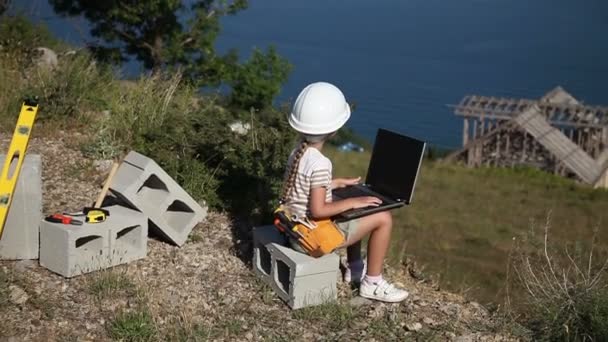 A criança é a construtora. Menina no capacete de construção e com óculos de sol. Ela está segurando laptop. Polegares para cima. Está bem. — Vídeo de Stock