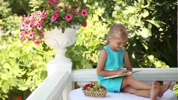 Petite fille souriante lisant un livre dans le jardin. Elle rit et montre Ok à la caméra. Le raisin est sur la table — Video