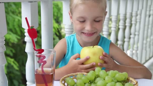 Chica comiendo manzana amarilla en la mesa. En la mesa hay una cesta de uvas y un vaso de jugo de manzana. Pulgares arriba. Ok. — Vídeos de Stock