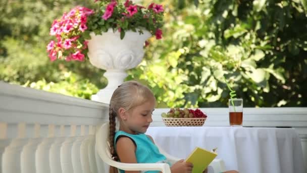Niña de preescolar leyendo un libro en el jardín. Niño sentado en una silla en la mesa. Sobre la mesa hay un vaso de jugo y una cesta de uvas. Ella abrazando el libro y mirando a la cámara — Vídeos de Stock