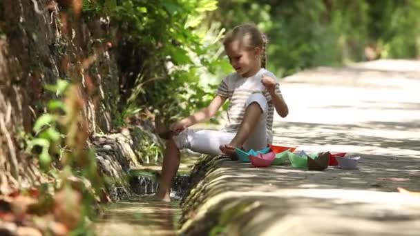 Chica sentada al lado de un arroyo y lanzando una gran cantidad de barcos de papel en el agua — Vídeos de Stock