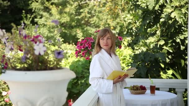 Junger Erwachsener im Bademantel auf der Veranda. Mädchen liest ein Buch und lächelt in die Kamera — Stockvideo