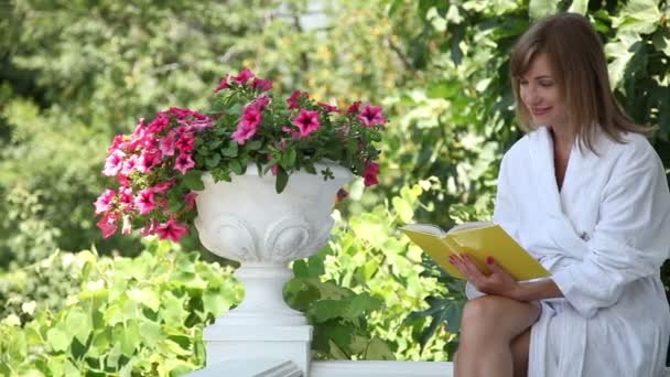 Jovem adulto relaxante no jardim. Uma mulher a ler um livro. Ela está vestida com um roupão de banho — Vídeo de Stock