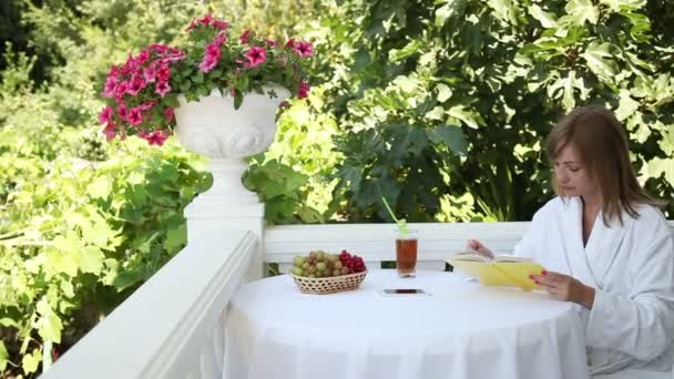 Junge Erwachsene lesen ein Buch und lächeln in die Kamera. die an einem Tisch sitzende Frau. Tisch steht auf dem Balkon im Garten — Stockvideo