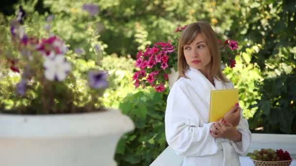 Mladý dospělý v županu stojící na přední verandě. Ona drží žlutá kniha — Stock video