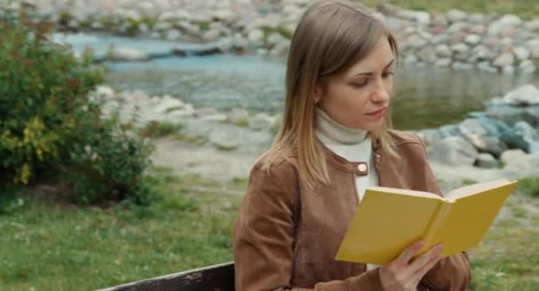 Ung vuxen kvinna läser en bok. Flickan sitter på en bänk i parken och ler mot kameran — Stockvideo