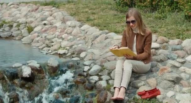 Портрет счастливой молодой взрослой женщины, отдыхающей в парке и читающей книгу. Девушка сидит на земле возле ручья — стоковое видео