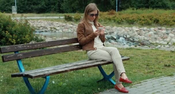 Mujer adulta joven usando el teléfono celular y sentada en el banco en el parque — Vídeo de stock