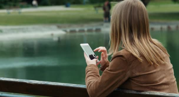 Портрет молодой взрослой женщины, сидящей на скамейке в парке и использующей мобильный телефон — стоковое видео