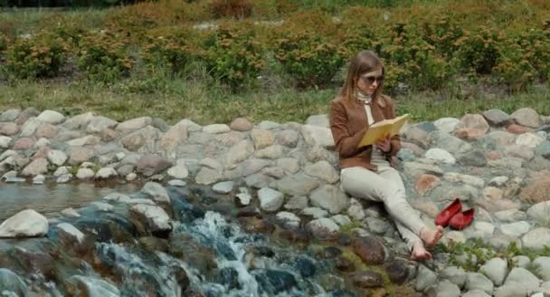 Mujer adulta joven descansando en el parque y leyendo un libro. Chica está sentada en el suelo cerca del arroyo — Vídeos de Stock