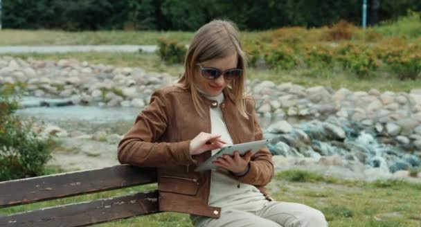 Mulher adulta jovem usando tablet pc e sentado no banco contra o pano de fundo do riacho — Vídeo de Stock