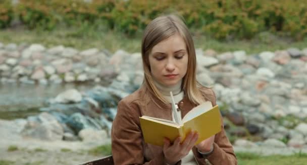 Retrato de mujer adulta joven leyendo un libro. Chica sentada en un banco en el parque y sonriendo a la cámara — Vídeos de Stock