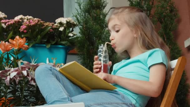 Rindo menina ler livro e beber suco e sorrindo para a câmera. Zumbido — Vídeo de Stock