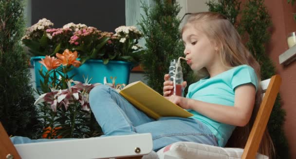 Śmiejąca się dziewczyna, czytanie książki i picie soku i uśmiecha się do kamery — Wideo stockowe