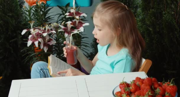 Mädchen liest ein Buch, trinkt Erdbeersaft und blickt in die Kamera — Stockvideo