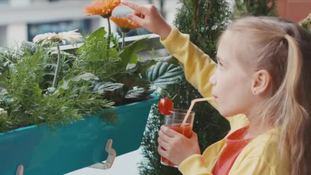 Blondes Mädchen trinkt Tomatensaft. Kind hält ein Glas Saft in der Hand und blickt auf Blumen und Kamera. Daumen hoch. Ok. zoomen — Stockvideo