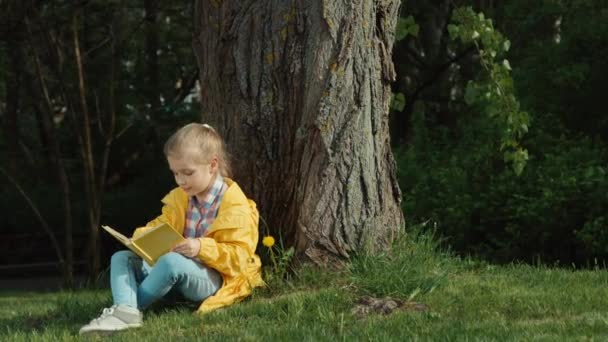 Ein Kind liest ein Buch, sitzt unter einem Baum, blickt in die Kamera und umarmt ein Buch. Zoomen — Stockvideo