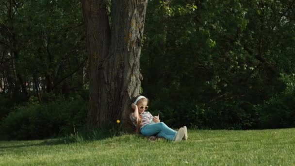 Mädchen hören Musik. Mädchen tanzt, während sie unter einem Baum liegt und in die Kamera blickt. Zoomen — Stockvideo