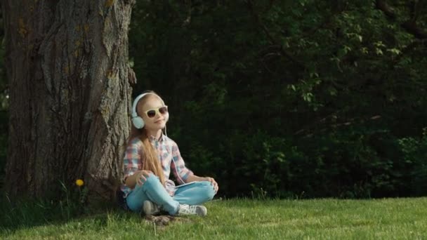 Chica escuchando música. La chica está bailando mientras está sentada bajo un árbol. Ampliación — Vídeos de Stock