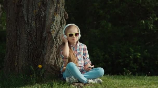 Дівчина слухає музику. Дитина сидить під деревом. У неї в руках телефон. Збільшення — стокове відео