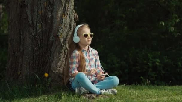 Ragazza che ride ascoltando musica. Bambino seduto sotto un albero. Nelle sue mani il telefono. Il bambino e 'online. Zoomare — Video Stock