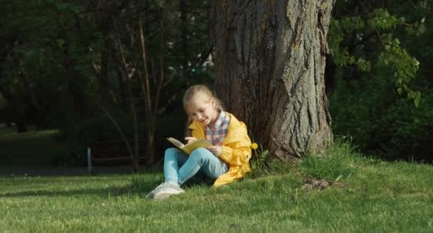 Bambino che legge un libro seduto sotto un albero e guarda la macchina fotografica. Pollice in alto. Va bene. — Video Stock
