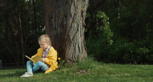 Niño leyendo un libro sentado debajo de un árbol y mirando a la cámara y abrazando un libro — Vídeo de stock