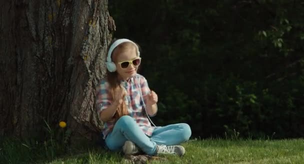 Chica escuchando música. Chica está bailando mientras se sienta debajo de un árbol y mirando a la cámara — Vídeos de Stock